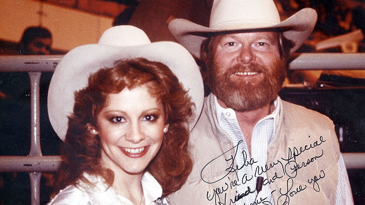 Reba McEntire and Red Steagall, both with red hair and wearing white cowboy hats, pose for a photo. The photo is autographed with a marker.